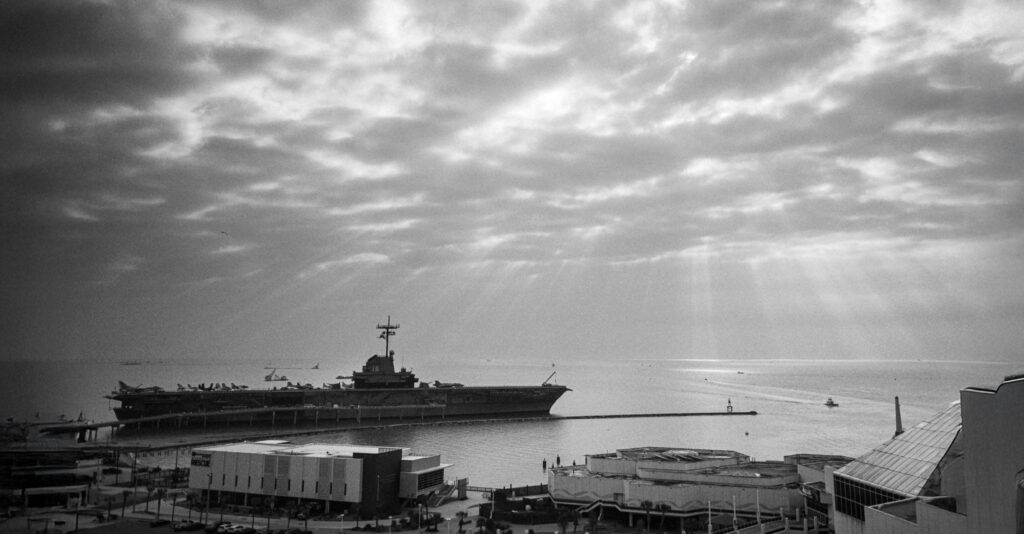 Black and white digital photograph of the USS Lexington aircraft carrier museum docked in Corpus Christi Bay, Texas, photographed from the waterfront under dramatic overcast skies. Documentary photography by Chris Marinez. #somewhereinTexas #LostInTexas